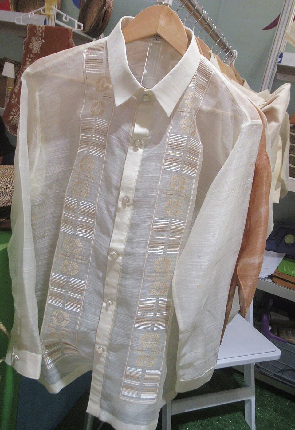 A barong tagalog hanging neatly on a wooden hanger near a window with natural light